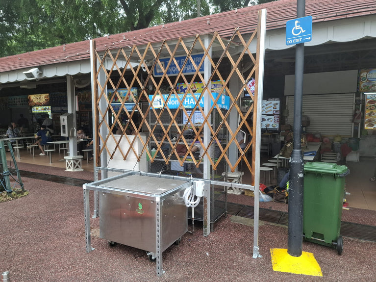 Misting pipework mounted on a wooden trellis beside a stainless steel equipment cabinet at an open-air food centre.