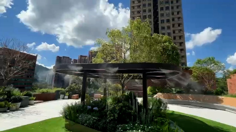 Wide view of mist cooling around a circular garden pergola with planting beds and surrounding buildings.